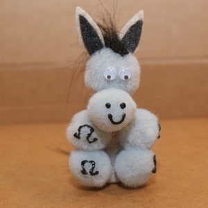 May include: A gray and black stuffed donkey toy with googly eyes and a smiling face. The donkey is made of pom-poms and has a black mane.