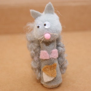 May include: A gray felt finger puppet shaped like a cat with pink ears, a pink nose, and large googly eyes. The cat is wearing a pink bow and has a brown felt tail.