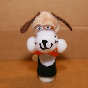 May include: A brown and white finger puppet dog wearing a black and orange bow tie. The dog has large white eyes and a black nose.