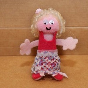 May include: A small felt doll with blonde hair, a pink flower, a red dress, and a patterned skirt. The doll has a smiling face with pink cheeks and black eyes.