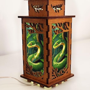 Op de afbeelding: Een houten lantaarn met een gedetailleerd ontwerp. De lantaarn heeft een rechthoekige vorm met uitgesneden bladerpatronen en een structuur met twee niveaus. De panelen tonen een groene slangenillustratie tegen een jungle-achtergrond. Een draad loopt vanaf de basis.