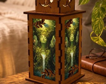 May include: A square wooden lantern with a light-up panel featuring a green-toned illustration of a figure with foliage hair and a waterfall. The wooden frame has laser-cut leaf details on the sides and a tiered top section with cutouts.