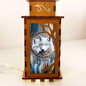 Op de afbeelding: Houten lantaarn met een gesneden ontwerp. Het voorpaneel toont een dromenvanger met twee witte wolven. De lantaarn heeft een warme gloed, ideaal voor het creëren van sfeer. Hij wordt gevoed door een snoer.