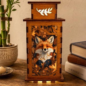 May include: A rectangular wooden lantern featuring a laser-cut design of a fox resting among autumn leaves. The top panel displays a cutout of a leafy branch. The lantern emits a warm internal glow, highlighting the orange and brown tones.