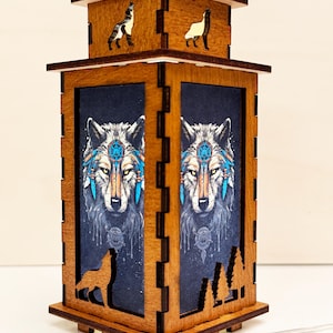 Op de afbeelding: Een houten lamp met een wolfontwerp. De lamp heeft een donkerblauwe achtergrond met een wolfsgezicht in het midden. De wolf draagt ​​een blauw en wit hoofdstel met veren. De lamp heeft een houten frame met uitsnijdingen van wolven boven en onder.