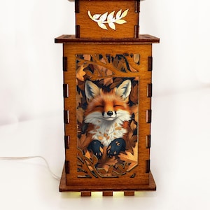 Könnte beinhalten: Holzlampe mit einer Fuchsdarstellung. Die rechteckige Lampe zeigt ein detailliertes Bild eines Fuchses inmitten von Herbstblättern. Die Oberseite der Lampe hat ein dekoratives Blattdesign. Die Lampe ist aus braunem Holz und strahlt warmes Licht aus.