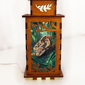 Op de afbeelding: Houten lamp met een rechthoekig frame en een gedetailleerde slangenillustratie. De lamp heeft een bruine afwerking met uitgesneden bladeren en een wit bladaccent aan de bovenkant. De slang is bruin en bruingeel, opgerold op een tak.