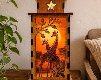 Op de afbeelding: Een rechthoekige houten lantaarn met een uitgesneden ster aan de bovenkant en een verlicht paneel met twee giraffen in een savannelandschap tijdens zonsondergang. De lamp heeft een donkerbruin frame met zichtbare verbindingen.