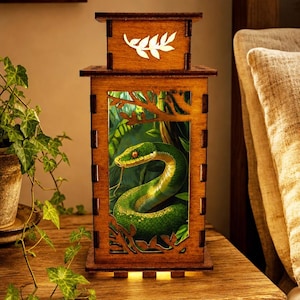 May include: A rectangular wooden lantern featuring a laser-cut frame with a top panel displaying a white leaf cutout. The central panel depicts a green snake coiled within a jungle scene, illuminated from within to highlight the vibrant colors.