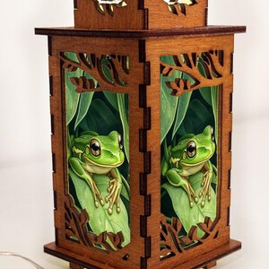 May include: A wooden lantern with a detailed frog and leaf design. The lantern is made of dark brown wood and features cut-out leaf shapes. Inside, a green frog sits on a leaf, creating a nature-themed decorative piece.
