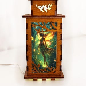 May include: A wooden fairy lamp with a rectangular shape. The lamp features a detailed illustration of a fairy in a forest scene, illuminated by an internal light. The lamp has a decorative leaf design on the top and is made of brown wood.