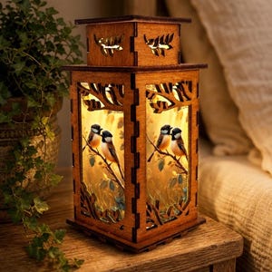 May include: A square wooden lantern with a warm internal glow, featuring laser-cut panels that depict pairs of small birds perched on branches. The top section has cutouts of leaves, and the structure is made of dark brown wood.