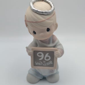 May include: A porcelain figurine of an angel with blonde hair and a silver halo, wearing a light blue and white robe. The angel holds a small rectangular chalkboard with the number 96 and the text Peace on Earth written in white.