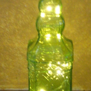 May include: A green glass bottle with a cork stopper, illuminated by small, warm white lights inside. The bottle has a decorative, textured surface and a classic shape, perfect for home decor.