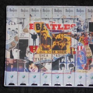 May include: A collection of eight VHS tapes featuring The Beatles, with a collage of images and text. The tapes are in a metal case, with the band's name and the word "Anthology" visible. The collage includes images from the band's performances and album covers.