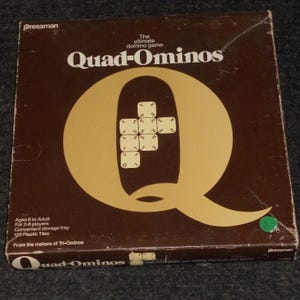 May include: A brown box for the game Quad-Ominos, with a large gold letter "Q" and the game's name in white. The box includes text indicating it's for ages 8 to adult, for 2-8 players, and contains 125 plastic tiles.