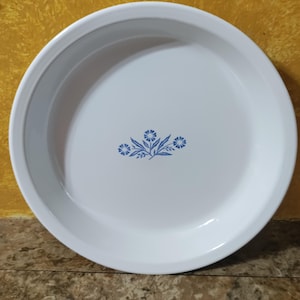 May include: A white, round serving dish with a blue floral design in the center. The dish has a slightly raised rim and a smooth, glossy surface. The floral design features stylized blue flowers and leaves.