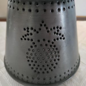 May include: A dark gray metal candle holder with a flared shape. The candle holder features a decorative pattern of punched holes in the shape of a pineapple. The metal has a slightly textured surface.