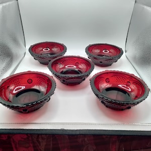 May include: A set of five small, round, ruby red glass bowls featuring a textured exterior pattern of raised dots and a diamond-like grid. Each bowl has a smooth, clear interior rim and a scalloped edge, arranged in a cluster on a white surface.
