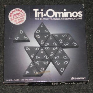 May include: Tri-Ominos board game box. The box is black with white text and triangular dominoes. The game is for 2 to 6 players, ages 8 to adult. The box says "Anniversary Edition" with larger, easy to read numbers.