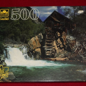 May include: A Golden Guild 500-piece jigsaw puzzle featuring a scenic landscape with a waterfall, a wooden structure, and lush greenery. The puzzle box has the words "FULLY INTER-LOCKING" printed on it.