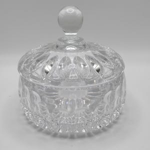 May include: A clear glass candy dish with a matching lid featuring a spherical knob handle. The bowl and lid are decorated with a pressed pattern of vertical ridges and scalloped edges, creating a reflective surface throughout the entire piece.