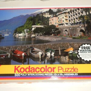 May include: A Kodacolor puzzle box featuring a scenic lake view with boats, buildings, and mountains. The puzzle contains 500 fully interlocking pieces and measures 13 x 19 inches when assembled. The box has a yellow, black, and red stripe design.