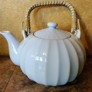 May include: White ceramic teapot with a ribbed body and a woven handle. The teapot has a lid with a small knob and a curved spout. The handle is made of woven light brown material. The teapot is designed for brewing tea.