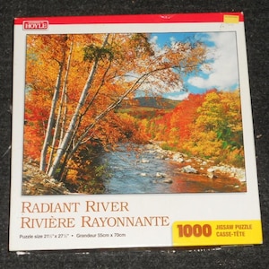 May include: A 1000-piece jigsaw puzzle featuring a vibrant autumn landscape with a river flowing through a forest of trees with orange and yellow foliage. The puzzle measures 21.5" x 27". The title is "Radiant River".