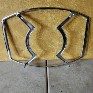 May include: A chrome-plated metal pot rack with a curved top and angled supports. The rack has a shiny, reflective surface and is designed to hold pots and pans. The supports have a curved design and are attached to the top frame.