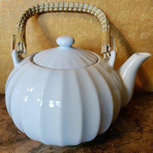 May include: A white ceramic teapot with a rounded, ribbed body and a matching lid. The teapot features a curved spout and a swing handle wrapped in light-colored woven rattan material, attached to two upright loops on the top of the vessel.