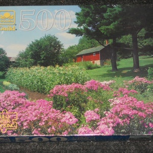 May include: A Golden Guild 500-piece jigsaw puzzle featuring a vibrant landscape scene. The image showcases a red barn, lush green trees, and a garden filled with pink flowers. The puzzle box has the words "Fully Interlocking".