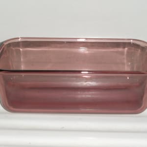May include: A rectangular, transparent, pink-tinted glass baking dish. The dish has a simple, classic design with slightly flared sides and small handles on each end. The glass appears smooth and clean, suitable for oven use.