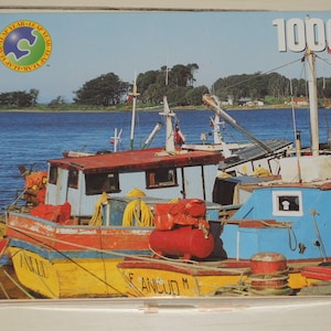 May include: A 1000-piece jigsaw puzzle featuring a coastal scene with colorful fishing boats. The boats are painted in red, yellow, and blue, with the word "ANCI" visible on one. The puzzle box has the "LEAP YEAR" logo.