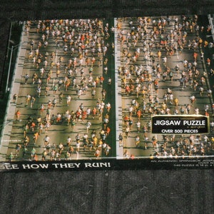 May include: A jigsaw puzzle featuring a bird's-eye view of a marathon with hundreds of runners on a road. The puzzle box has text that reads "JIGSAW PUZZLE OVER 500 PIECES" and "SEE HOW THEY RUN!" The puzzle measures 18 x 23.5 inches.