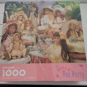 May include: A 1000-piece jigsaw puzzle featuring a tea party scene with antique dolls. The puzzle box is pink and has the text "the DOLLS: Tea Party" and "springbok by Hallmark". The assembled size is 24 x 30 inches.