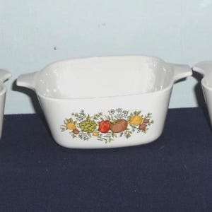 May include: Three white ceramic baking dishes with handles. The center dish features a colorful floral and vegetable design. The dishes are rectangular and appear to be vintage kitchenware.