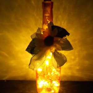 May include: A decorative glass bottle with a black stopper, filled with warm yellow fairy lights. The bottle is adorned with a sunflower and a checkered ribbon. The overall effect is a warm, inviting glow, perfect for home decor.