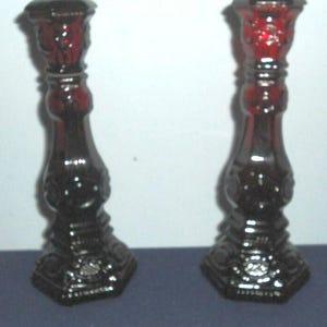 May include: Two ruby red glass candlesticks with ornate detailing. The candlesticks have a hexagonal base and a flared top. The design includes floral and scrollwork patterns. The glass has a glossy finish, reflecting light. The candlesticks are a matching pair.