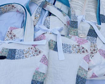 May include: Several handmade quilted tote bags with floral and patchwork designs. The bags have white quilted bodies, colorful patchwork accents, and blue velvet-lined handles. White ribbon bows add a decorative touch. A book is visible in the background.