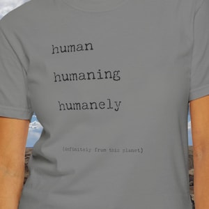 May include: A gray t-shirt with the text "human humaning humanely (infinitely from this planet)" printed on the front. The shirt is being worn by a person with dark hair and a neutral expression. The background is a cloudy sky and a rocky landscape.