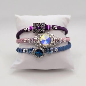 Könnte beinhalten: Drei übereinanderliegende Armbänder mit verschiedenen Stofffarben und Schmuckakzenten. Das obere Armband ist lila mit einem quadratischen Schmuckstein, das mittlere Armband ist rosa mit einem runden, schillernden Schmuckstein und das untere Armband ist blau mit einem runden blauen Schmuckstein.