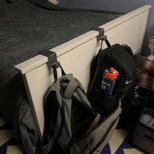 May include: A white bed frame with two dark gray metal hooks holding backpacks and a lunch bag. A black backpack with a Gatorade water bottle is visible. The bed is covered with a gray blanket, and a patterned armchair is in the background.