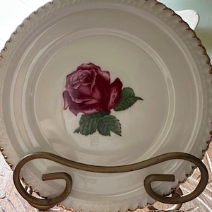 Pode incluir: Um prato de cerâmica branca com uma ilustração central de uma rosa vermelho-escura com folhas verdes. O prato tem uma borda recortada com um acabamento dourado. Está exposto num suporte de arame metálico curvo de cor bronze.