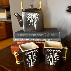 May include: A black ceramic teapot and two matching mugs with white bamboo designs. The teapot has a woven handle and a white leaf design. The mugs have bamboo handles. The set is displayed on a wooden table.
