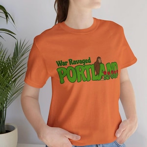 Peut inclure: T-shirt orange avec l'inscription "War Ravaged PORTLAND" en vert, représentant un Bigfoot de dessin animé et des roses rouges. Le t-shirt est à col rond et à manches courtes.