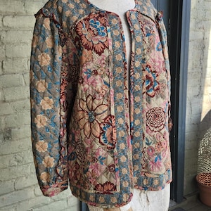 May include: A quilted jacket with a floral pattern in shades of pink, blue, and brown. The jacket is displayed on a mannequin and features a round neckline and open front. The fabric has a quilted texture.