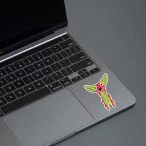 May include: A colorful sticker featuring a whimsical creature with large yellow ears, a red face, and a green and yellow body. The sticker is affixed to the trackpad of a laptop. The laptop is gray and the background is a solid gray.