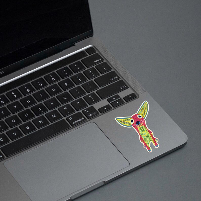 May include: A colorful sticker featuring a whimsical creature with large yellow ears, a red face, and a green and yellow body. The sticker is affixed to the trackpad of a laptop. The laptop is gray and the background is a solid gray.