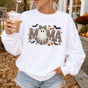 Peut inclure: Un sweat-shirt blanc à manches longues avec une impression graphique du mot MAMA en motif léopard. La lettre A est remplacée par un fantôme portant un nœud léopard. Le motif comprend des chauves-souris noires, des citrouilles et des étoiles.